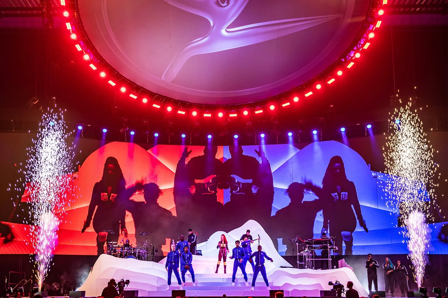 A stage performance with multiple performers, dancers, and musicians. The stage has a white, snowy mountain-like structure with large digital screens in the background showing silhouettes of performers. Fireworks are exploding on either side of the stage, and bright red, blue, and purple lights illuminate the scene.
