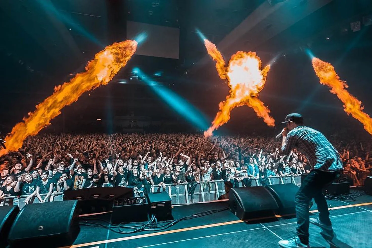 A performer in a baseball cap and plaid shirt on stage with a microphone, facing a large crowd at a concert, with flames shooting into the air from overhead pyrotechnics.