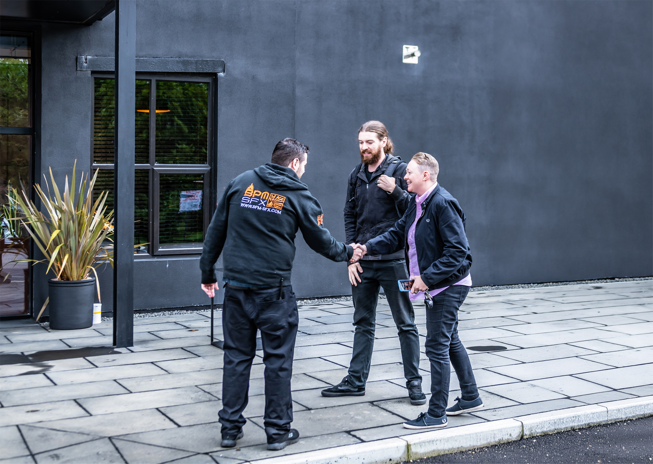 Three men standing outside on a paved sidewalk, shaking hands and smiling. One man is wearing a black jacket with orange writing on the back, another has a black backpack and beard, the third is holding a smartphone, and they are beside a gray building with windows and a large potted plant.