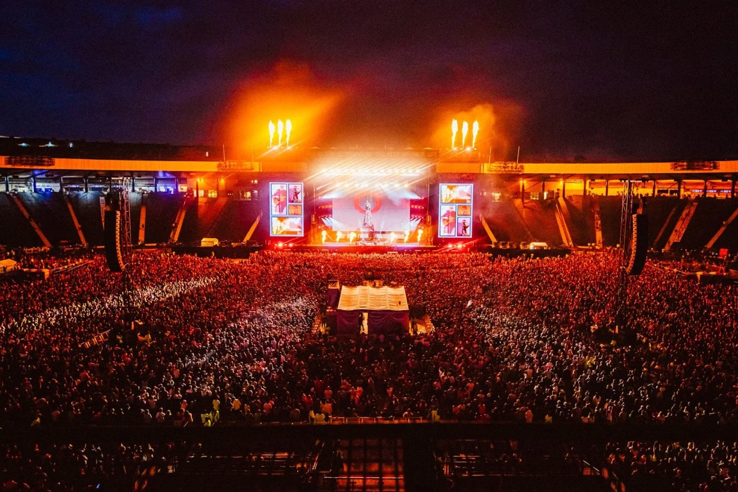 A large outdoor concert stage illuminated with bright lights and flames in the background at night, surrounded by a packed crowd at a stadium.