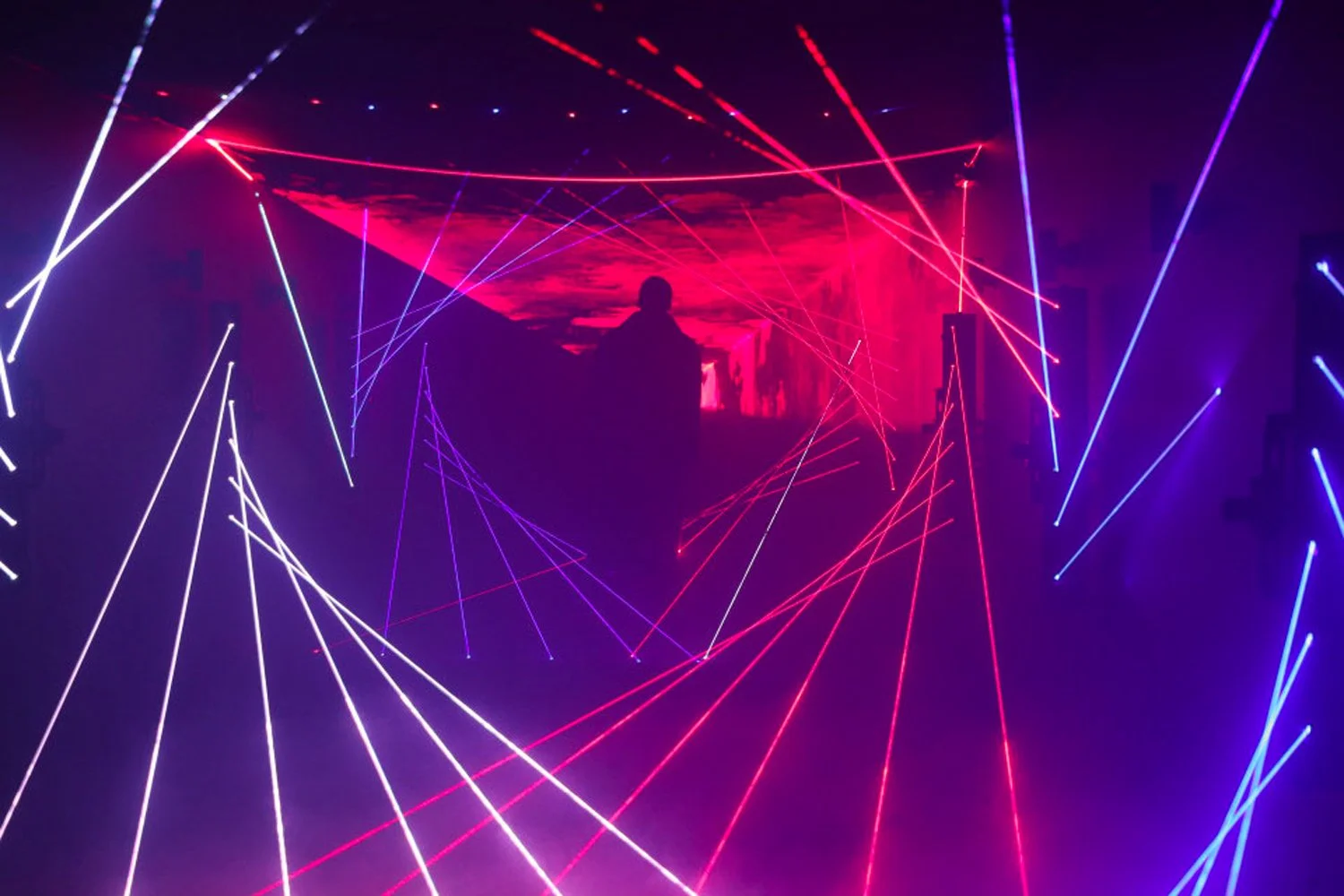Silhouette of a person standing in a dark room illuminated by a colorful laser light display, with red and purple laser beams creating geometric patterns across the space.