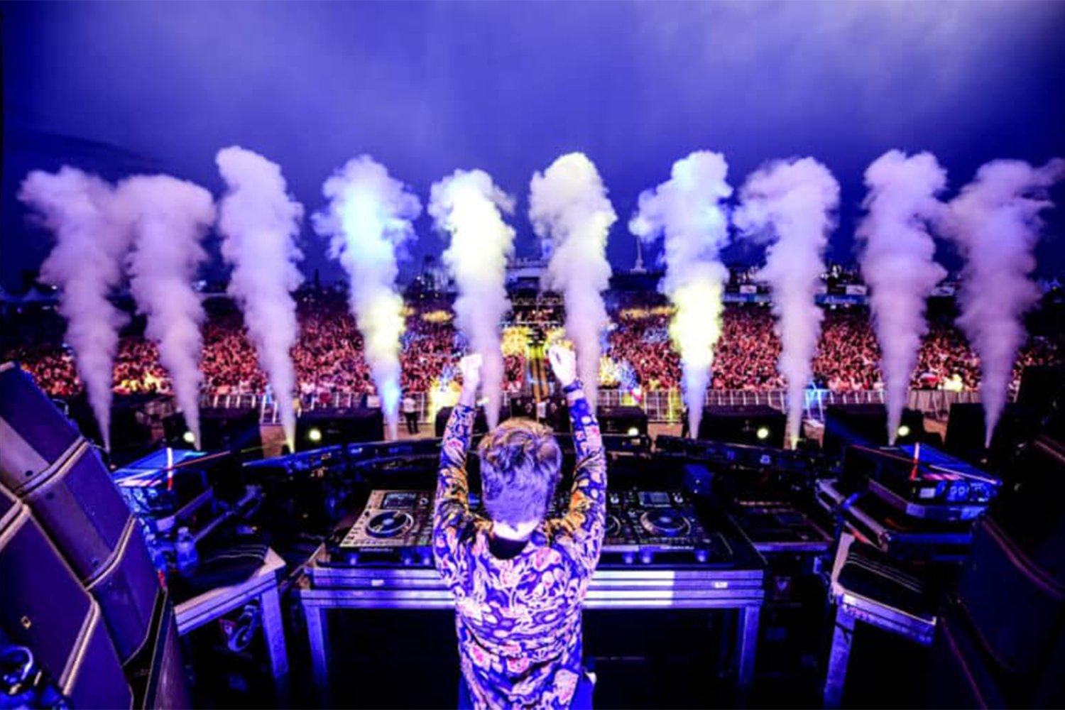 DJ performing at a concert with smoke effects, overlooking a large crowd at night.