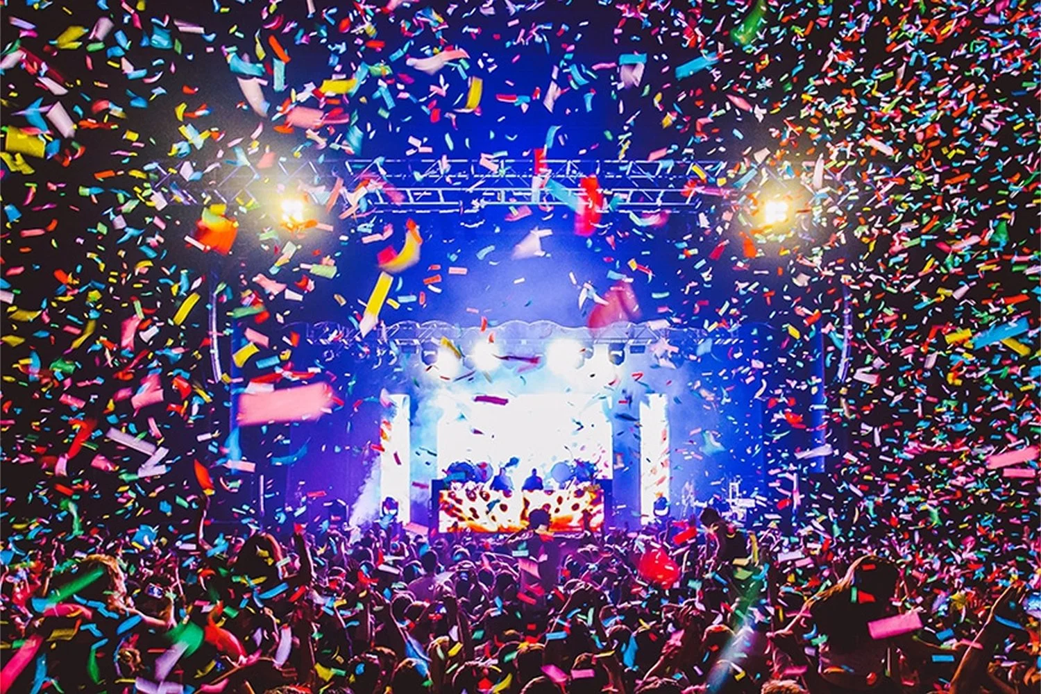 Crowd at a concert with colorful confetti falling, stage with bright lights and two DJs performing.
