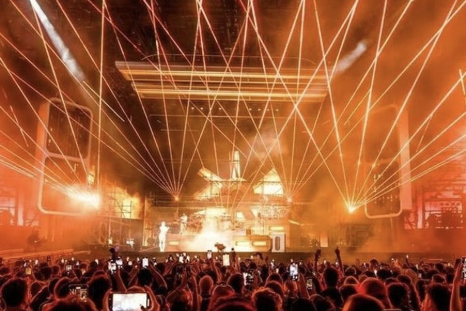 A concert scene with a large crowd of people taking photos or videos with their phones, bright orange and yellow stage lights, laser beams, and a smoke-filled atmosphere.
