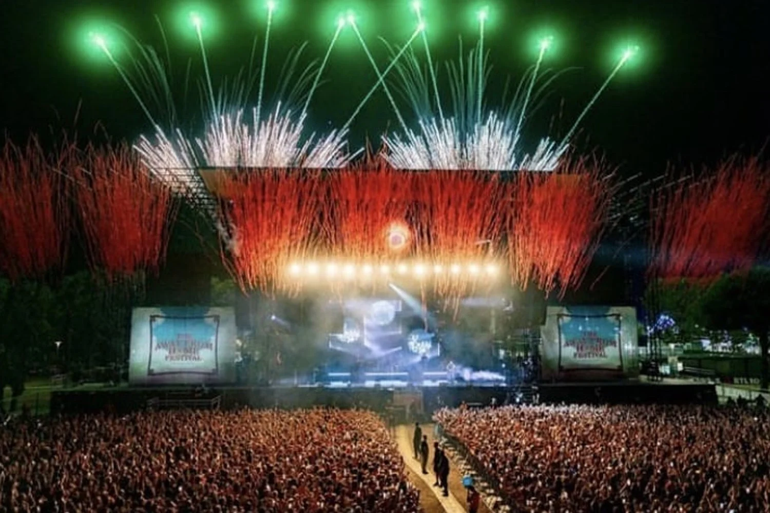 A large crowd gathered at an outdoor music festival during nighttime, with a stage illuminated and fireworks exploding in the sky above, displaying green, white, and red bursts.