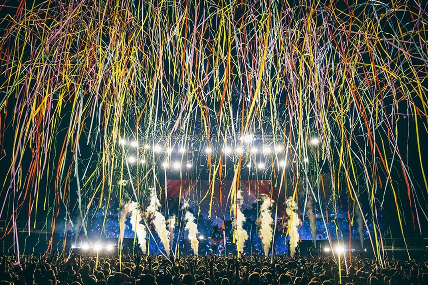Crowd at a concert with colorful confetti and fireworks in the air.