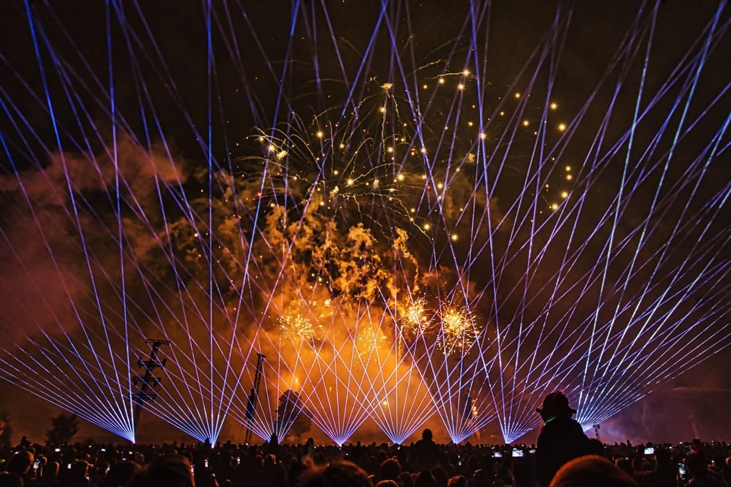A fireworks display at night with colorful explosions and laser-like light beams in blue and yellow, crowd watching from below.