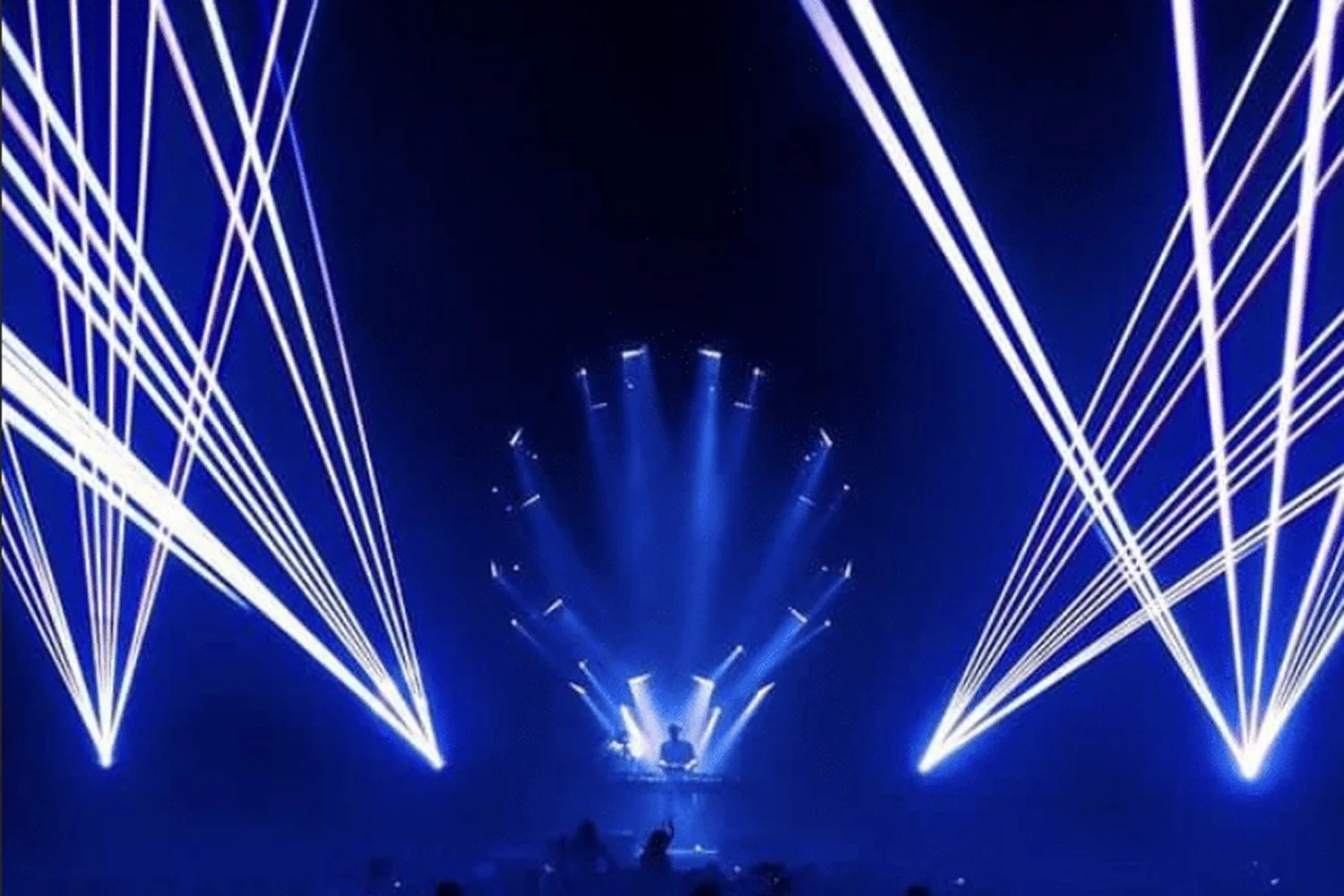 A dark stage with a DJ performing in front of an elaborate laser light show, featuring blue and white laser beams creating intricate patterns around the DJ.