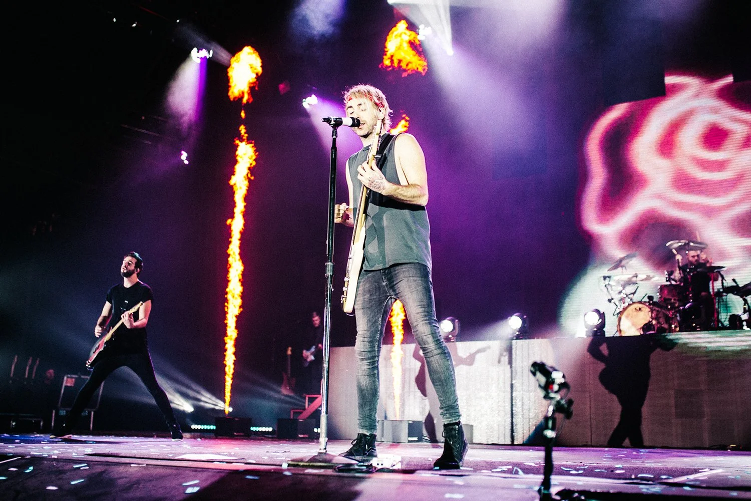 A live concert with a lead singer playing guitar and singing into a microphone, a bassist, and a drummer on stage, with colorful stage lights and flames in the background.