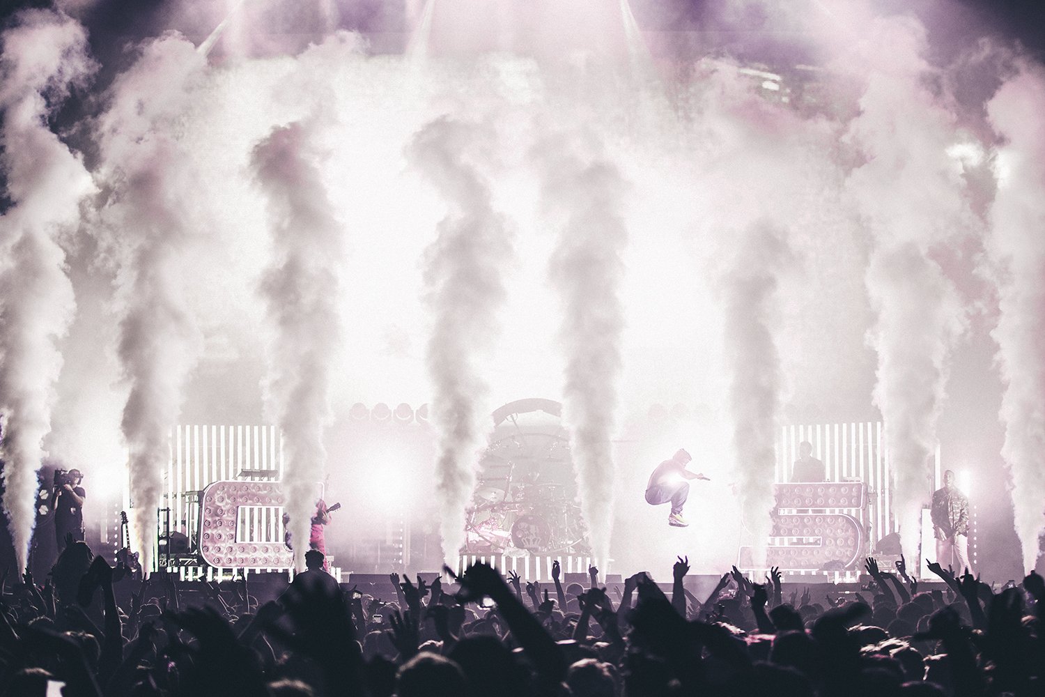 A concert scene with a large crowd, smoke effects, bright stage lights, and musicians performing with a singer jumping mid-air.