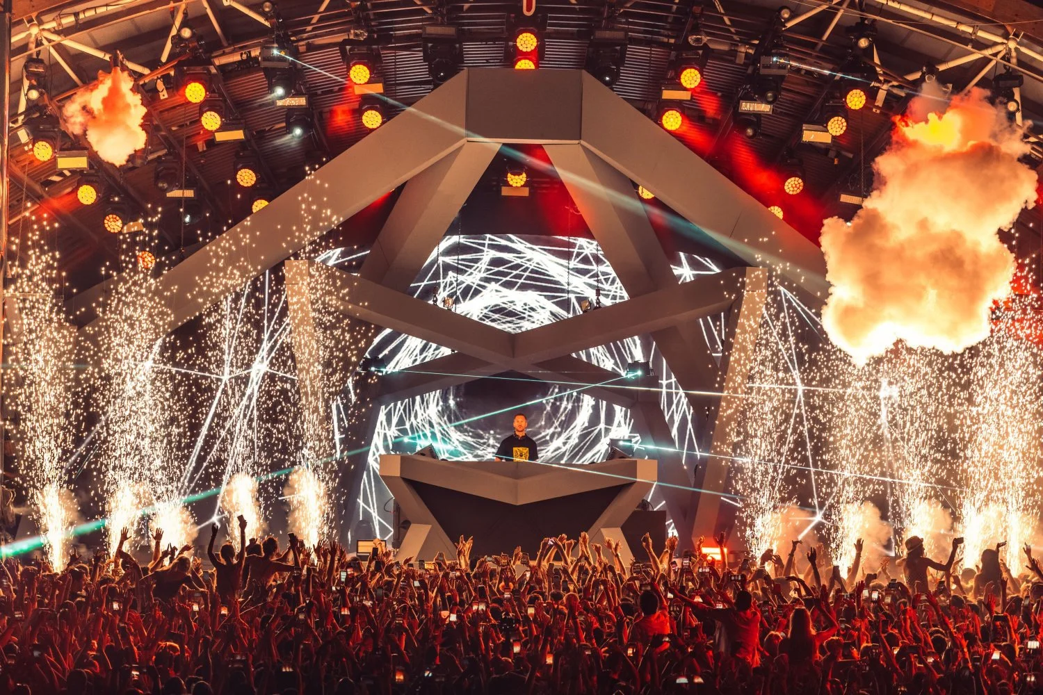 A lively indoor concert or festival with a DJ performing on stage, surrounded by pyrotechnics, fireworks, and bright lights, with a large crowd of people dancing and cheering.