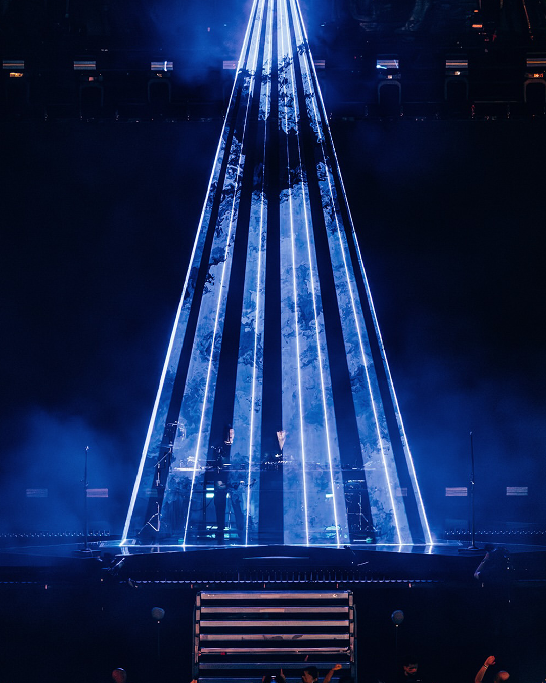 A concert stage with blue laser light beams creating a cone shape, with performers and musical equipment visible through the beams.