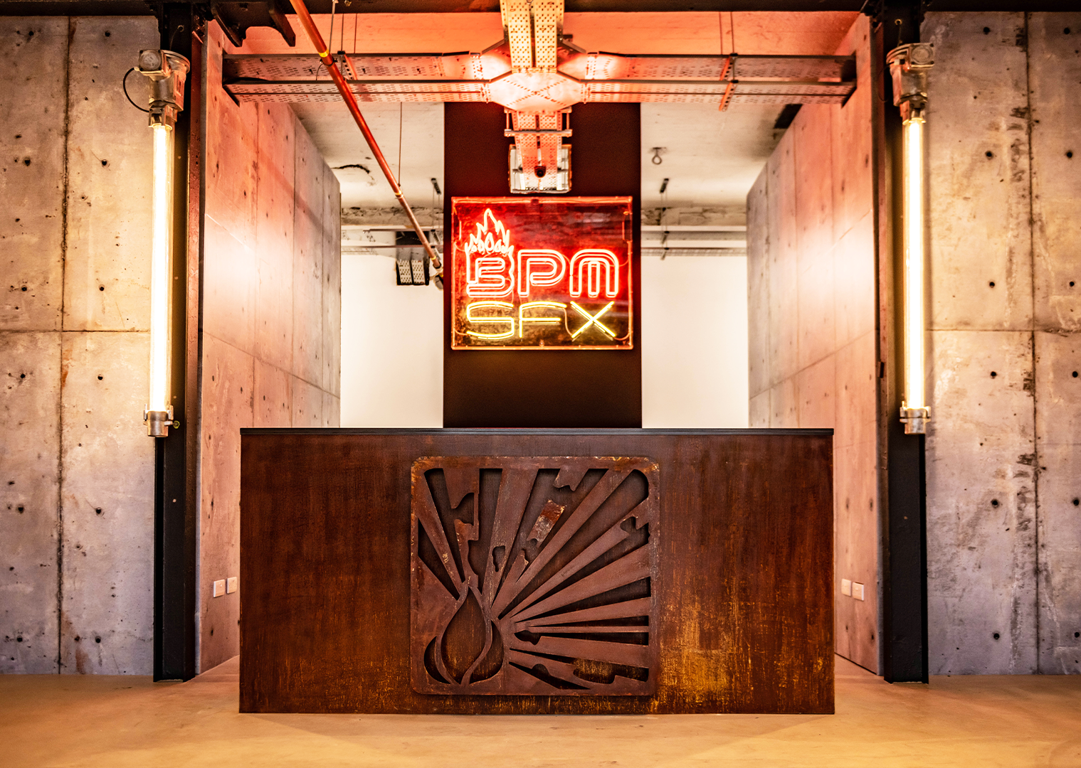 A modern office reception desk with a metal sign featuring a flame and vapor logo. In the background, a neon sign reads 'BPM SAX' in red and yellow. The setting has an industrial style with concrete walls, exposed pipes, and warm lighting.