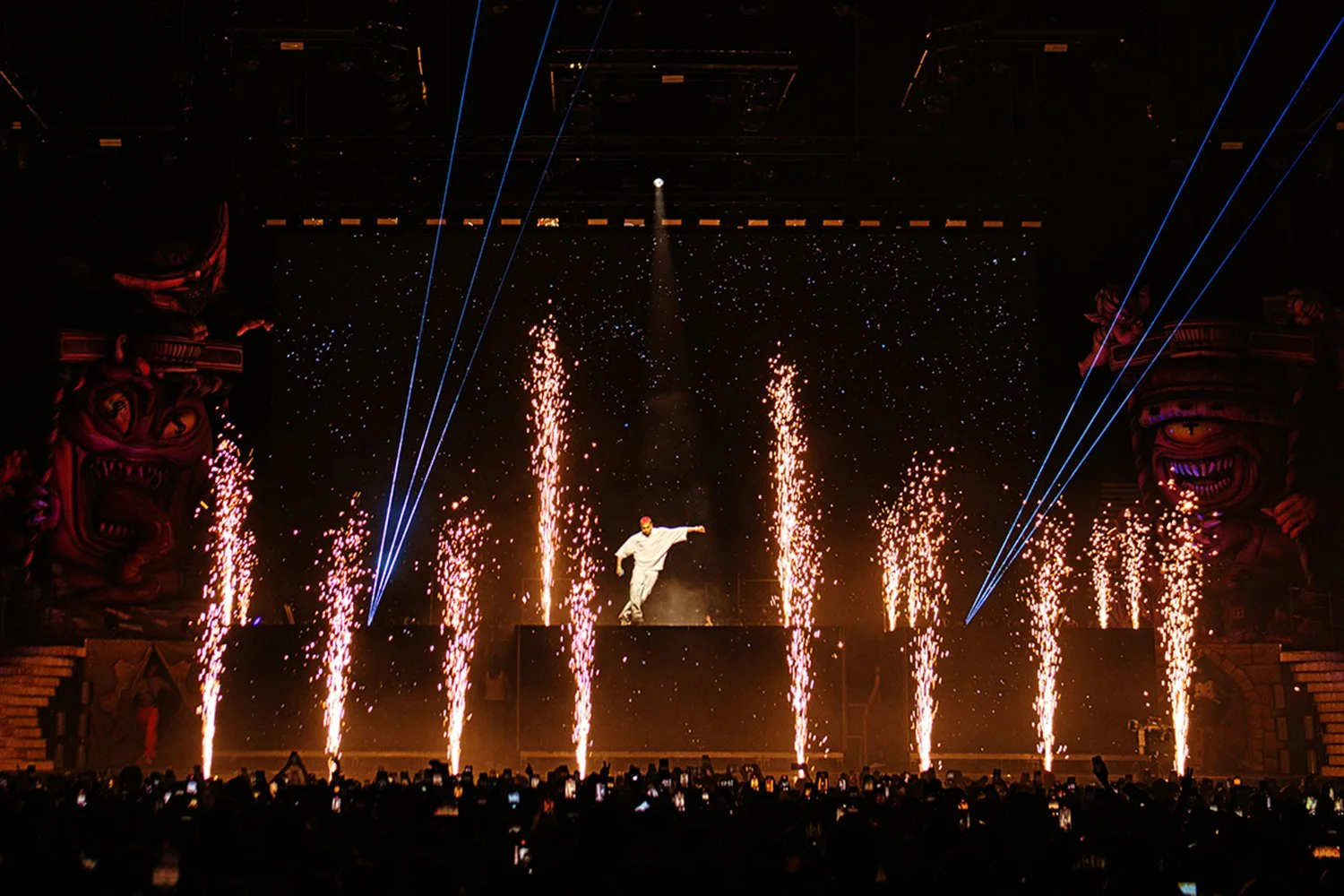 A performer dancing on stage with pyrotechnics and fireworks, surrounded by visual effects and large decorative statues.