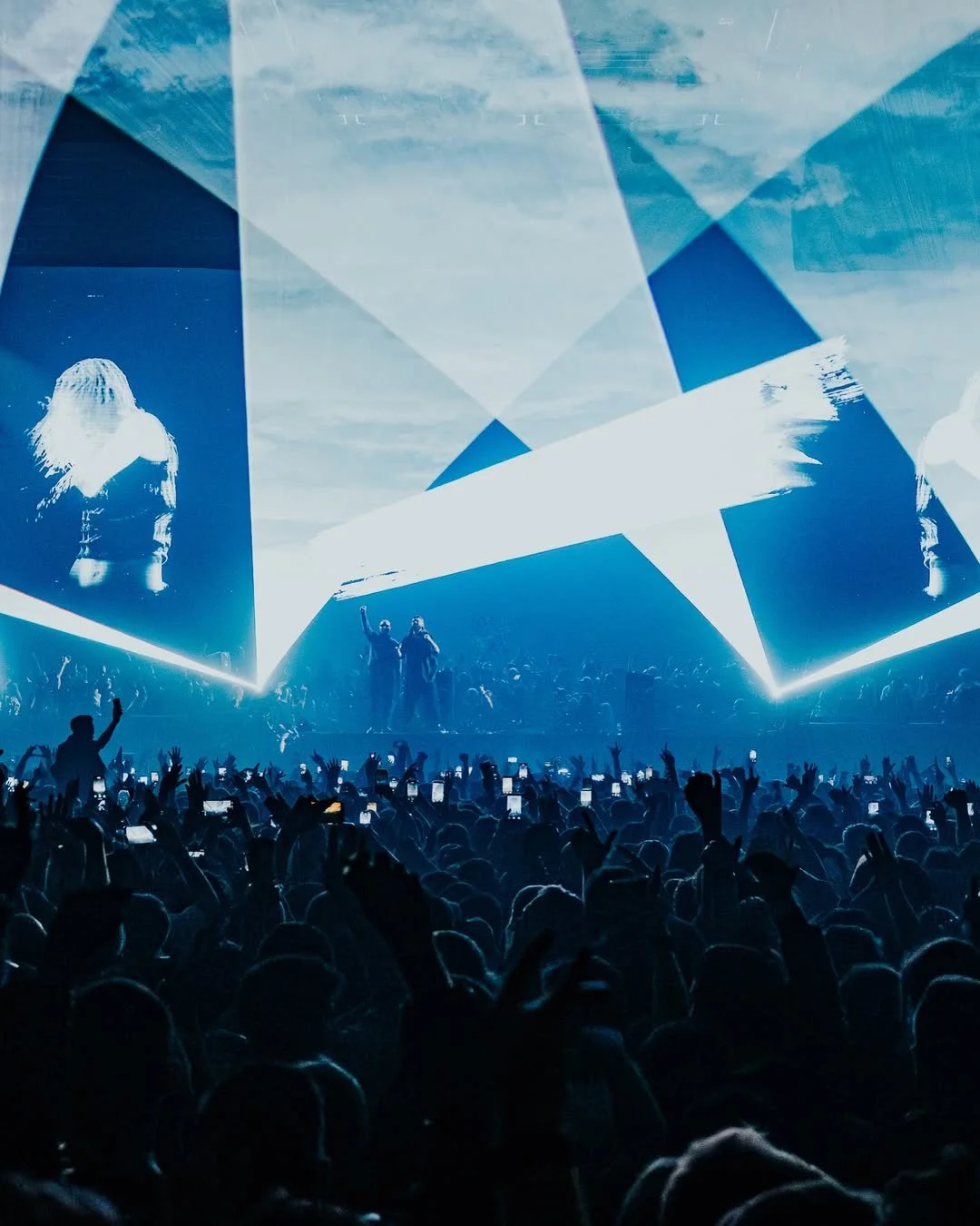 Crowd at a concert or festival with two performers on stage, large screens, and laser lights.