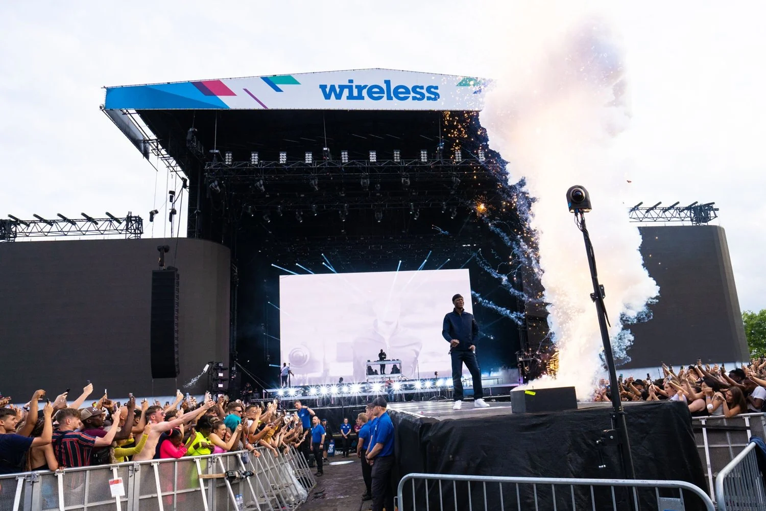 A large outdoor concert stage with a performer standing in front of a big screen, surrounded by cheering crowd, with fireworks and smoke effects, and a sign that says "wireless" above the stage.