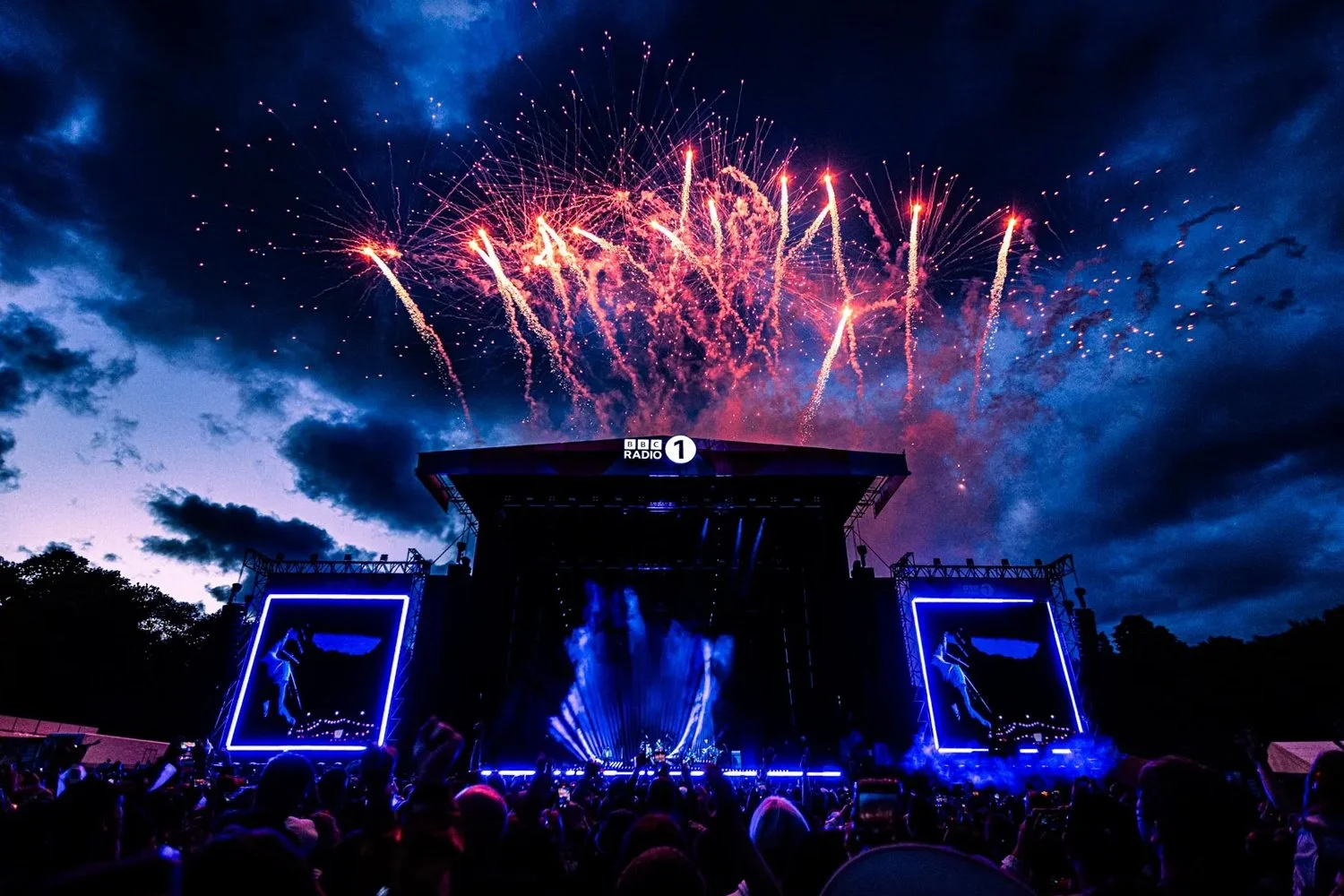 Fireworks exploding above a concert stage with a crowd in the foreground, BBC Radio 1 logo visible on the stage.