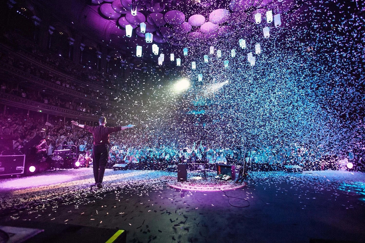 A performer standing on stage with arms outstretched amid a shower of confetti at a concert or event with colorful lighting and a large crowd watching.