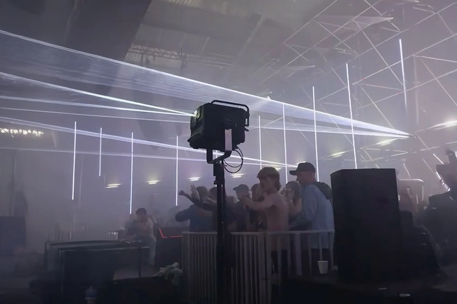 People dancing at an electronic music concert with laser lights and fog, a DJ is visible in the background.