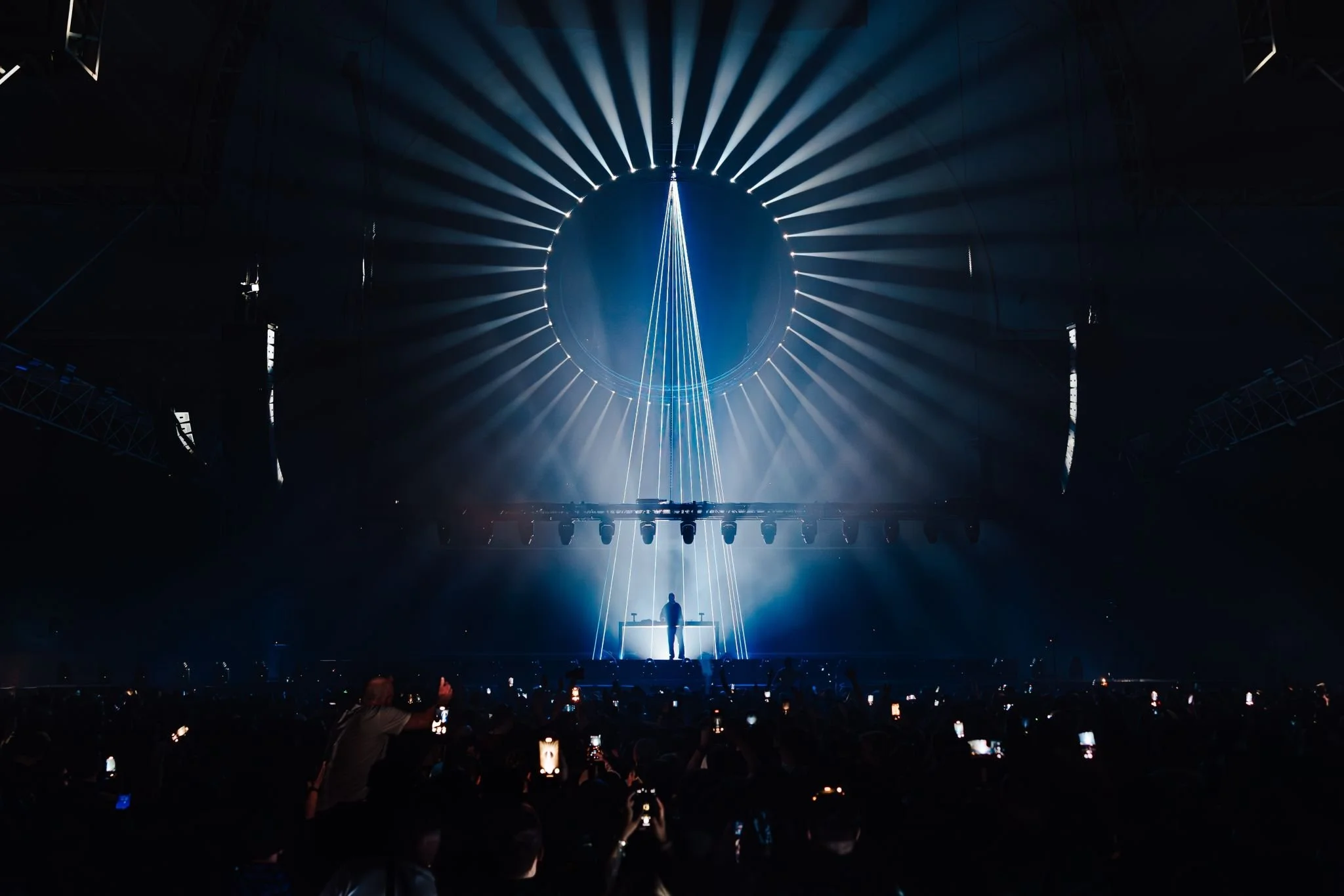 A person standing on a stage with a background of circular and vertical light beams, creating a dramatic lighting effect in a dark indoor venue, with audience members holding up phones.
