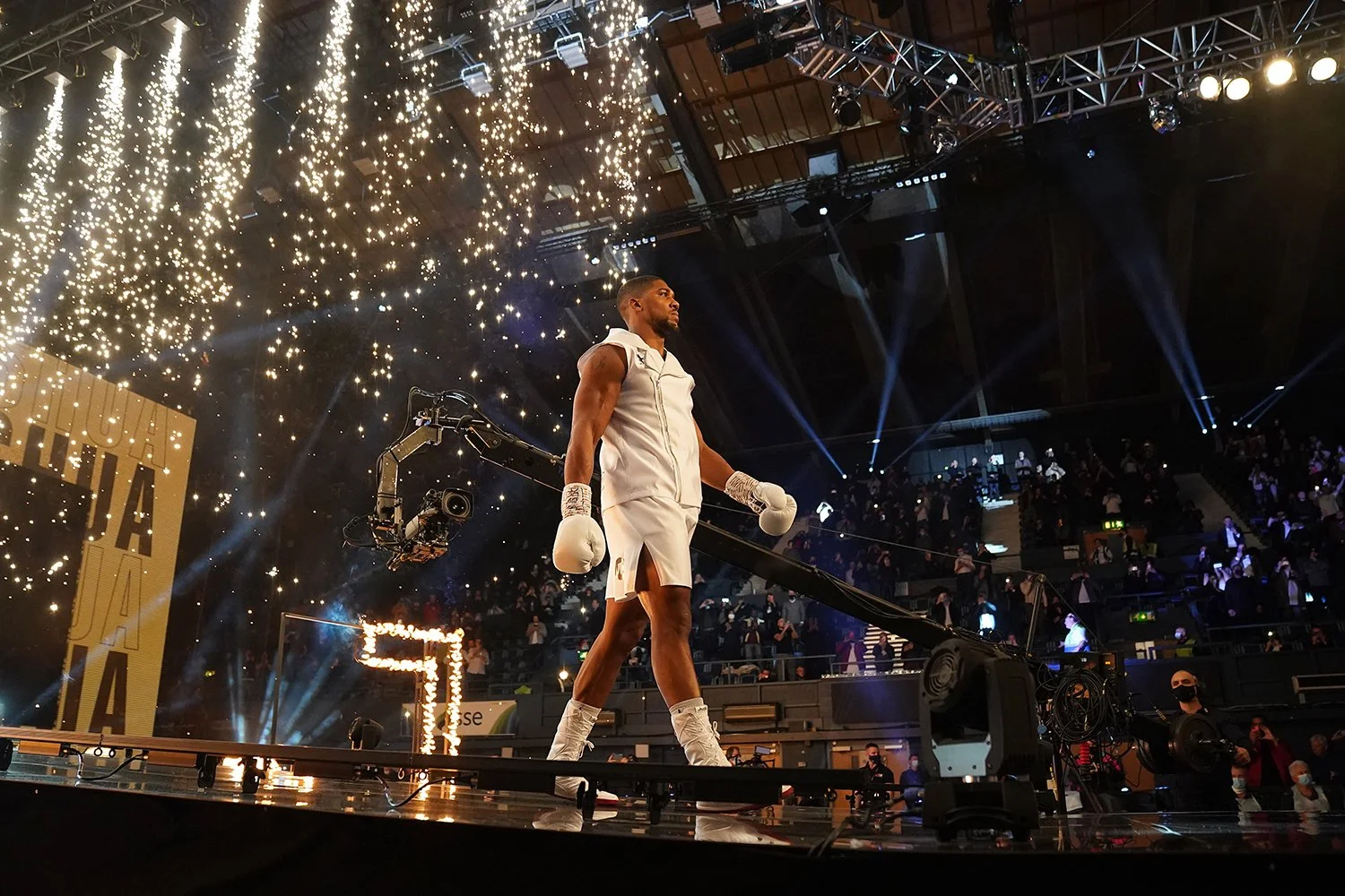 A boxer dressed in white in a boxing ring with sparklers and spotlights in the background during a live event.