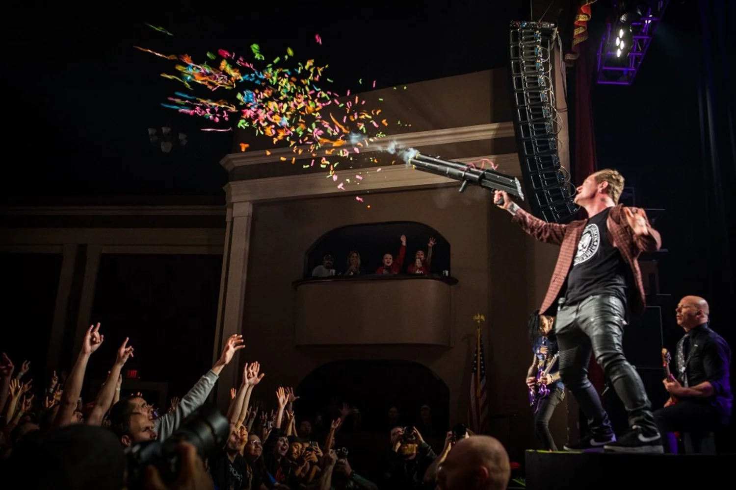 A performer on stage shooting a confetti gun into the crowd, with the audience reaching up and cheering. People observe from a balcony above.