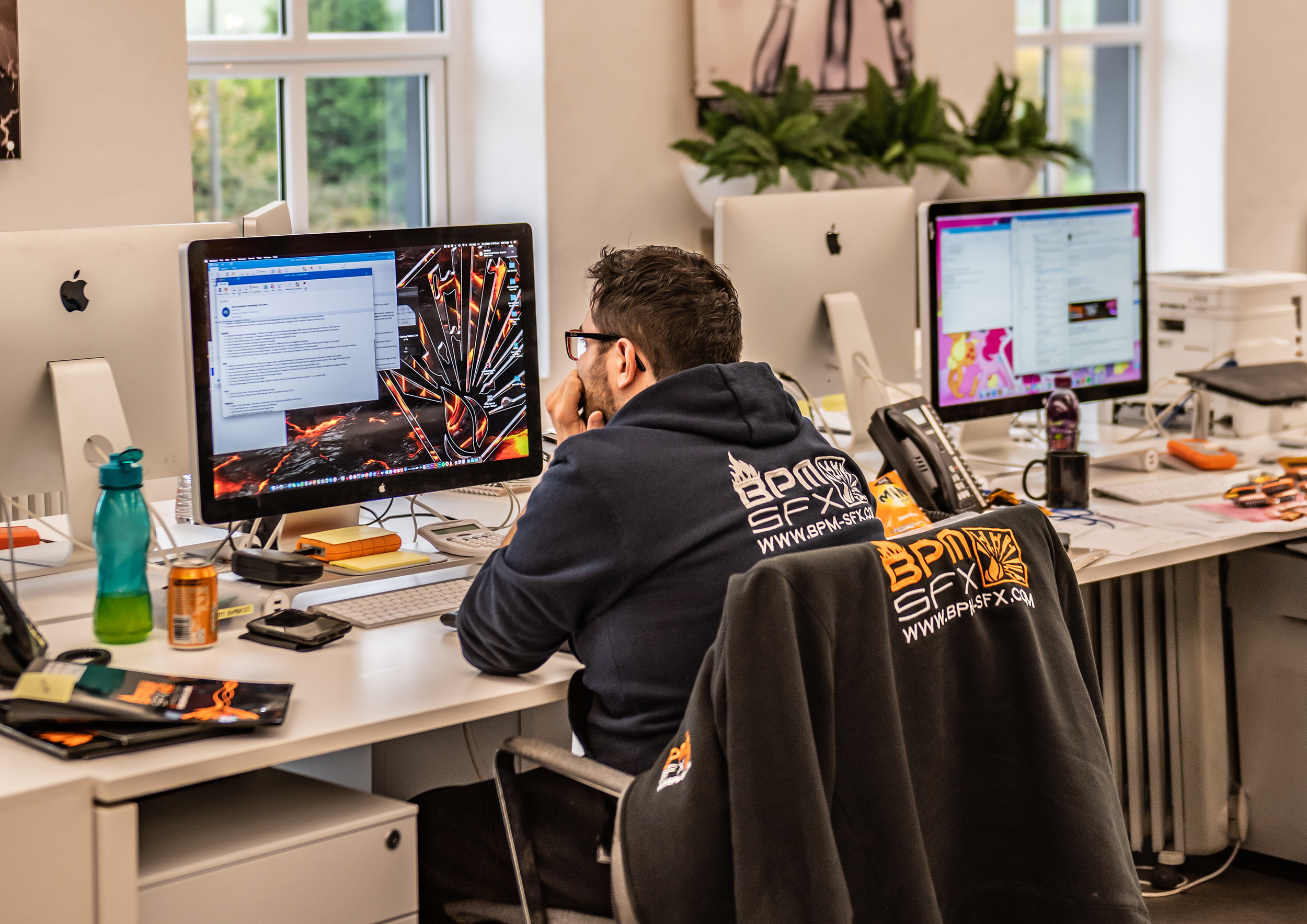 A man with glasses, wearing a black hoodie with printing on the back, is sitting at a desk in front of a computer. The desk has a water bottle, a couple of cans, a phone, and some papers. The room has large windows and potted plants in the background.