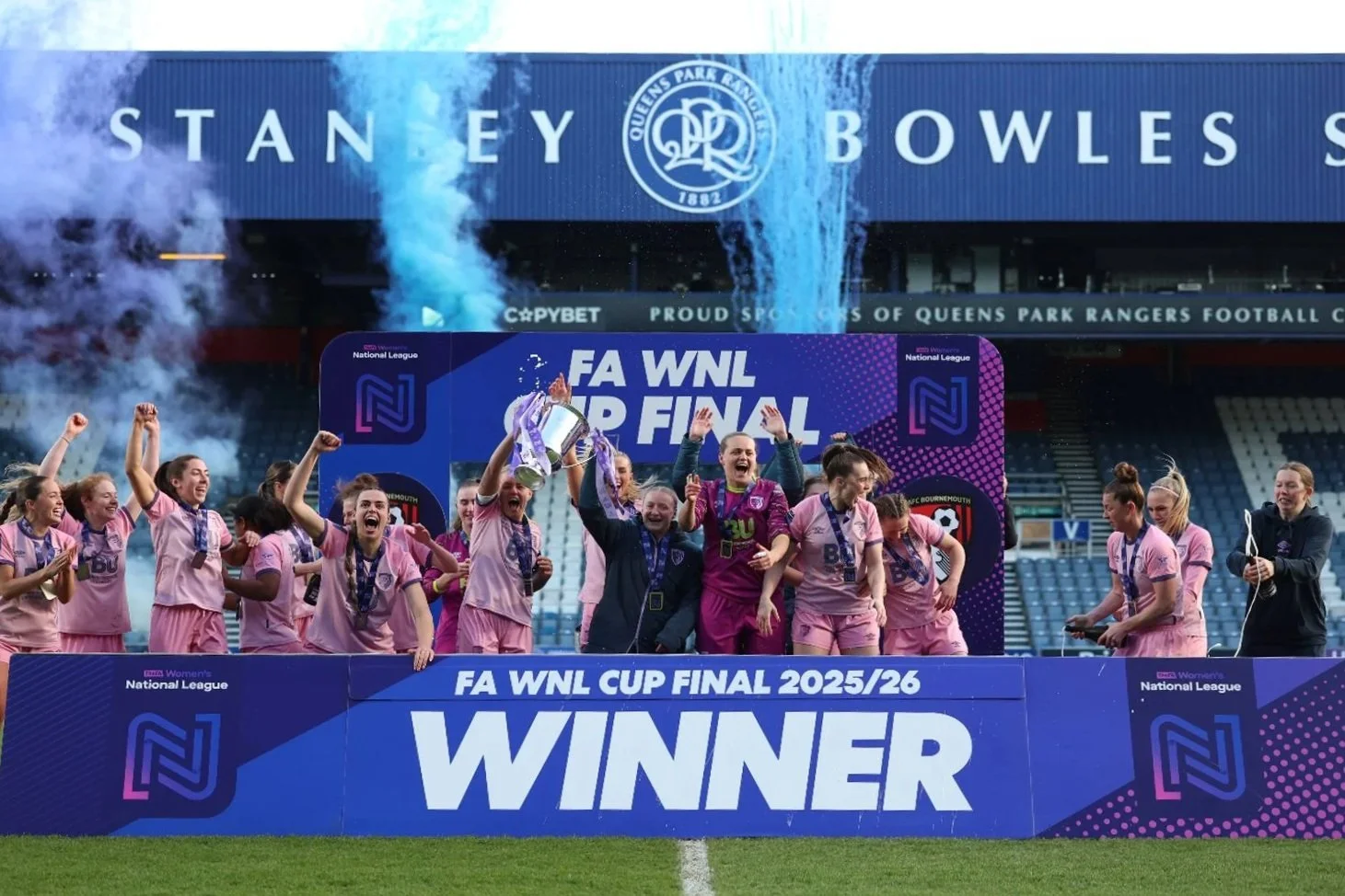 A group of women soccer players celebrating on the field after winning the FA WNL Cup Final 2025/26, with some holding a trophy, in front of a large blue and pink sign that says "WINNER" and sponsor banners, at a stadium with Queens Park Rangers branding.