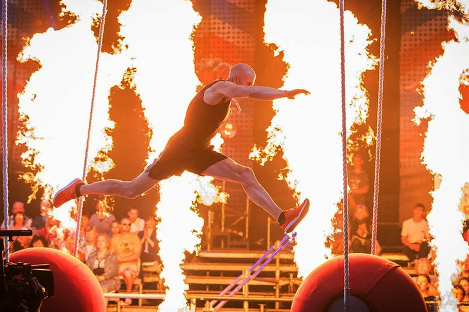A muscular man with a shaved head running on a platform between flames in a fire performance show, audience watching in the background.