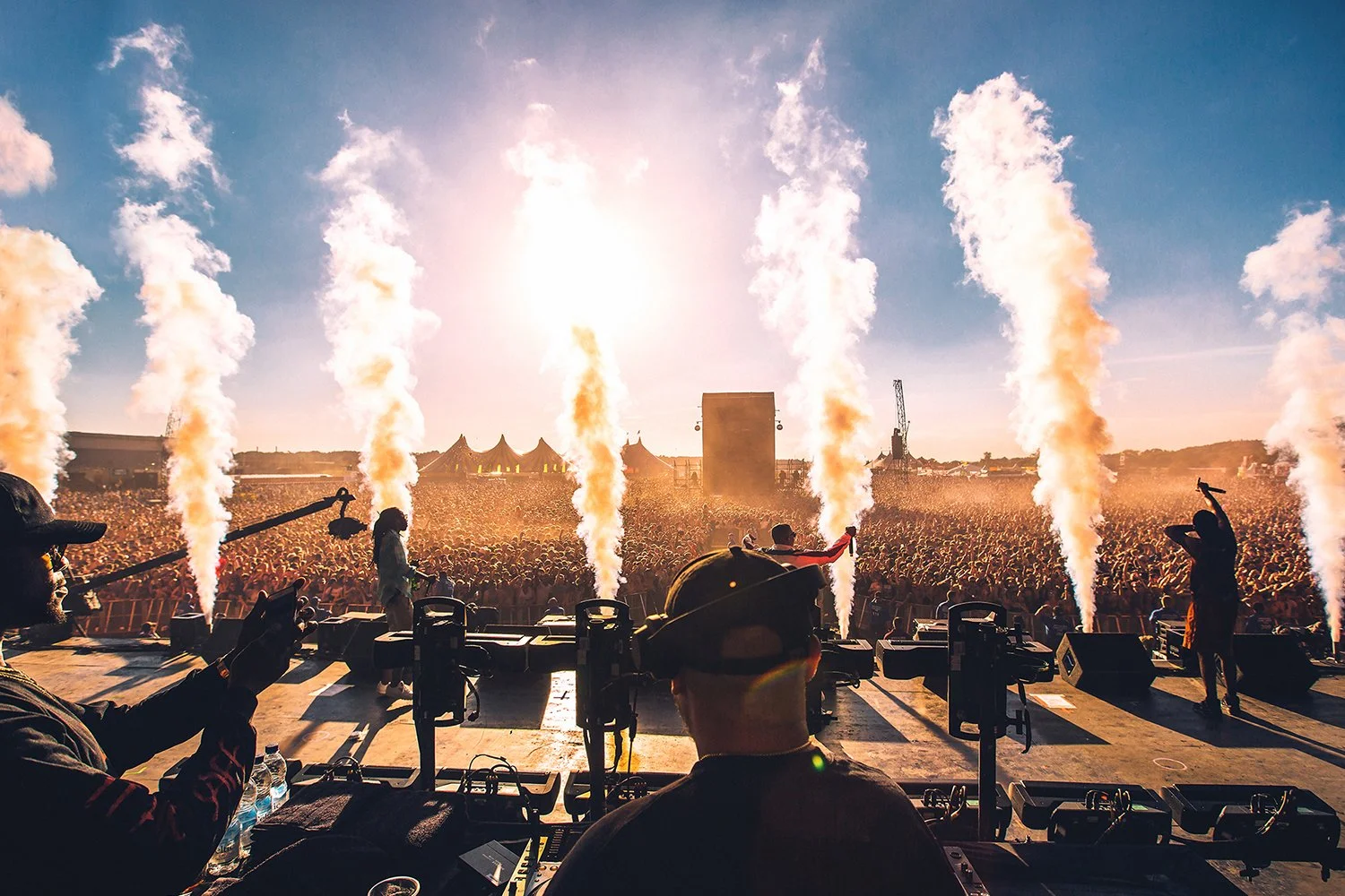 View from stage at outdoor music festival showing crowd, performers, and pyrotechnics with smoke blasts against the sun and clear sky.