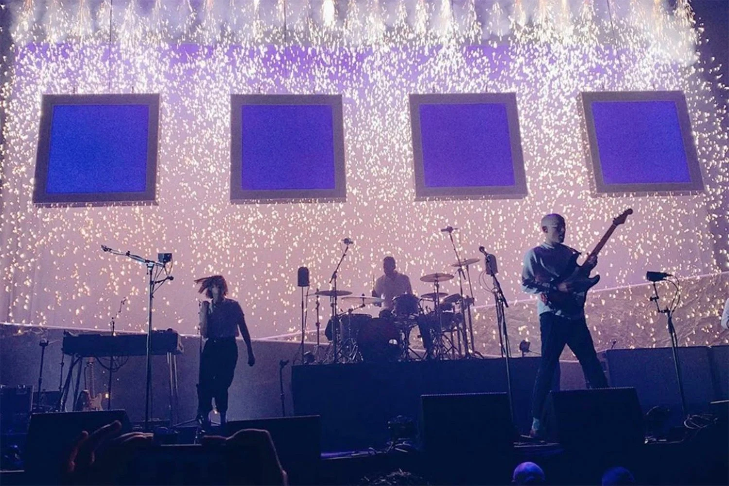 A band performing on stage with a visual display of falling sparks or fireworks behind them, including a keyboard player, drummer, and guitarist.