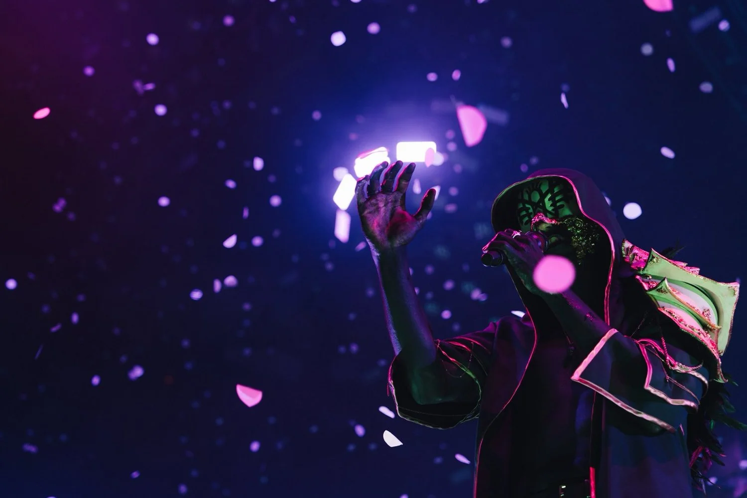 A performer on stage wearing futuristic clothing with neon accents, holding a microphone and raising their hand, with purple and pink confetti falling around them, against a dark background.