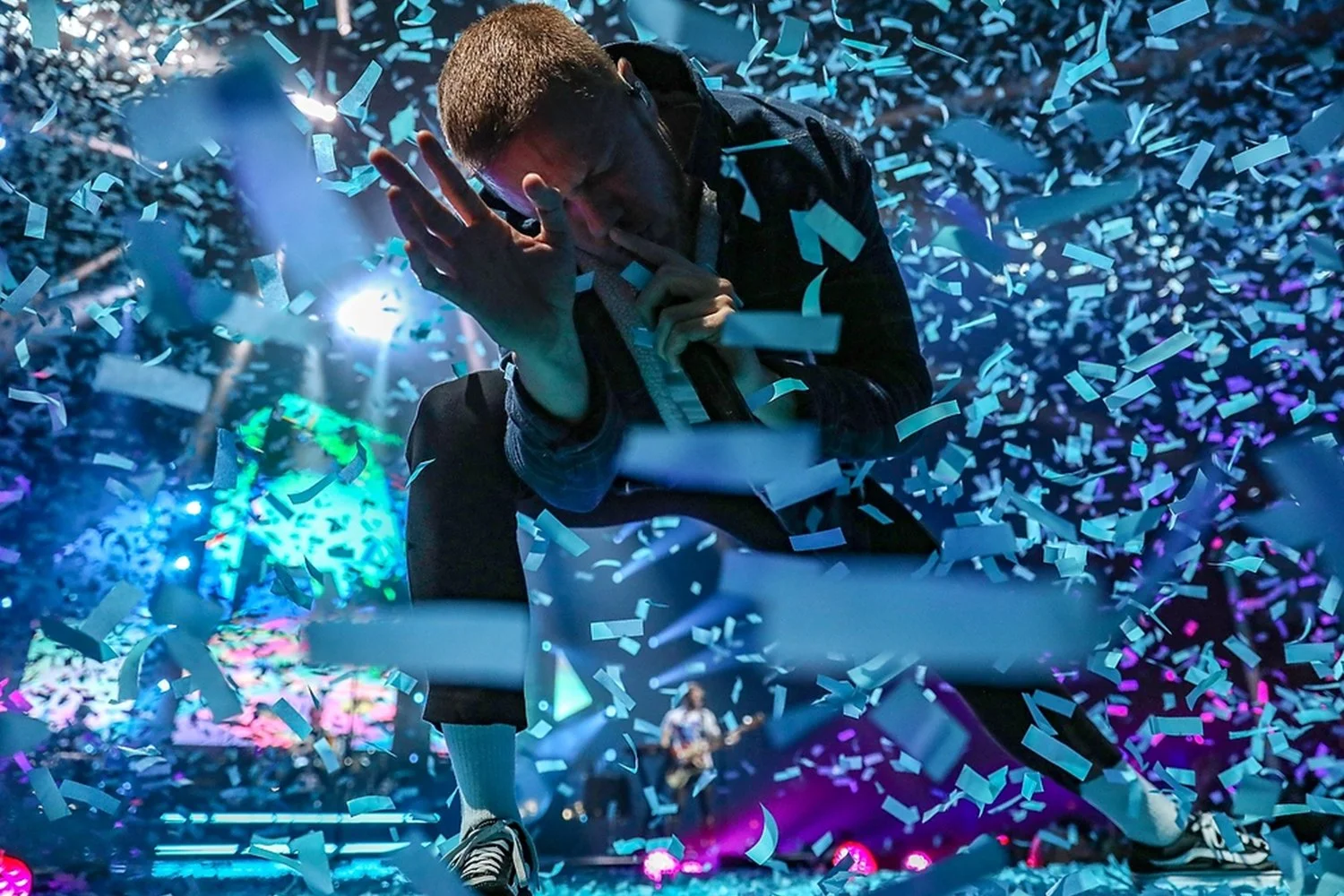 Performer singing on stage amidst colorful confetti with a person playing guitar in the background.