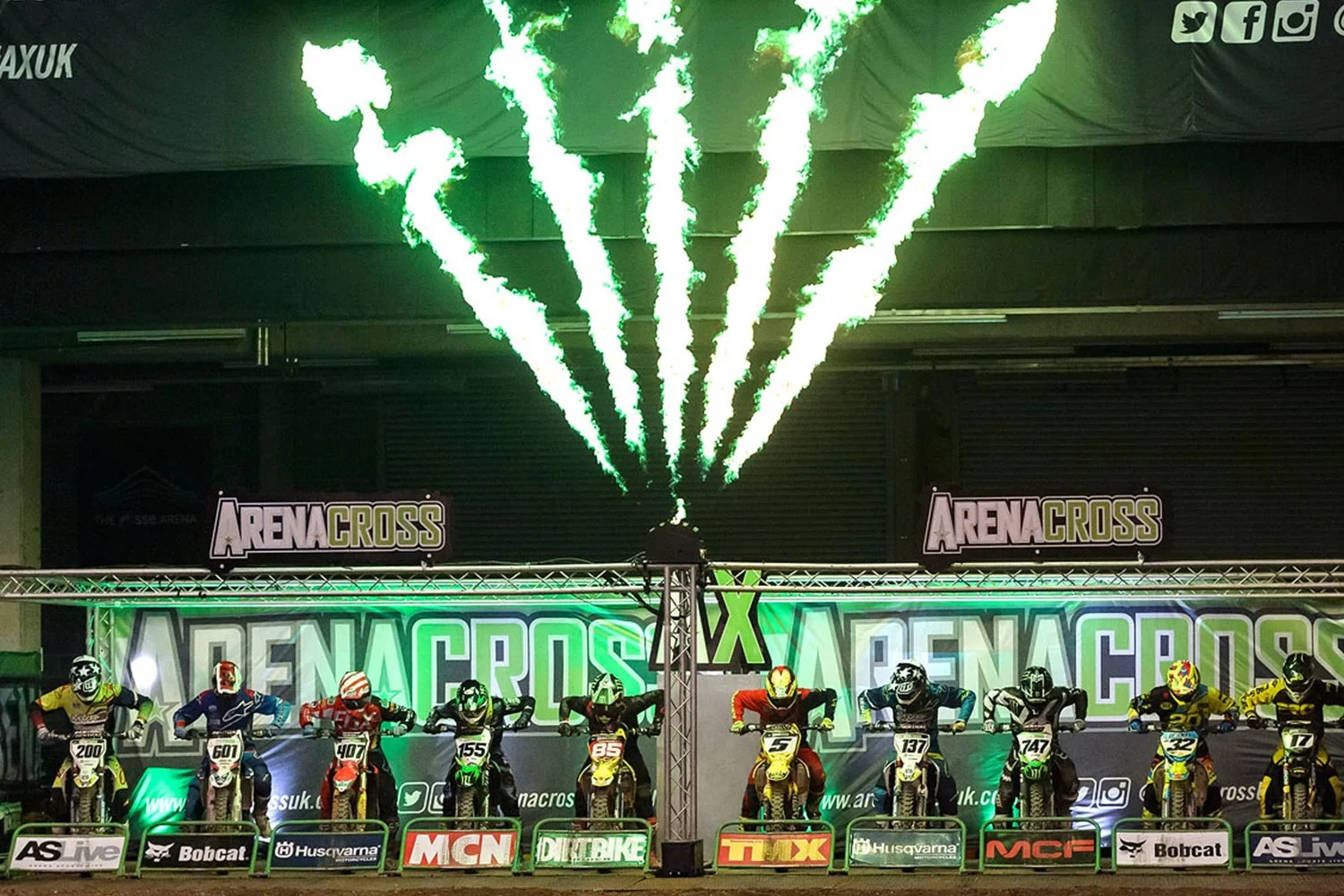 Motocross racers lined up on their bikes at a starting line, with a green fireworks display above them, in an indoor arena at an ArenaCross event.