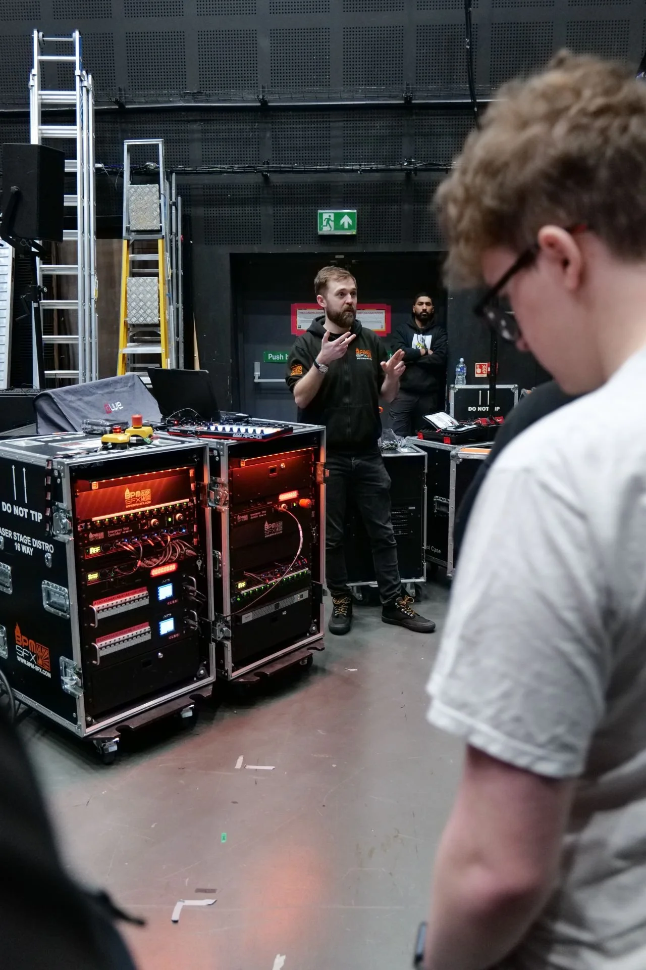 A man with a beard, wearing a black hoodie, is talking and gesturing to a group of people in a room with technical equipment, including servers, wires, and ladders, suggesting a technical or training environment.