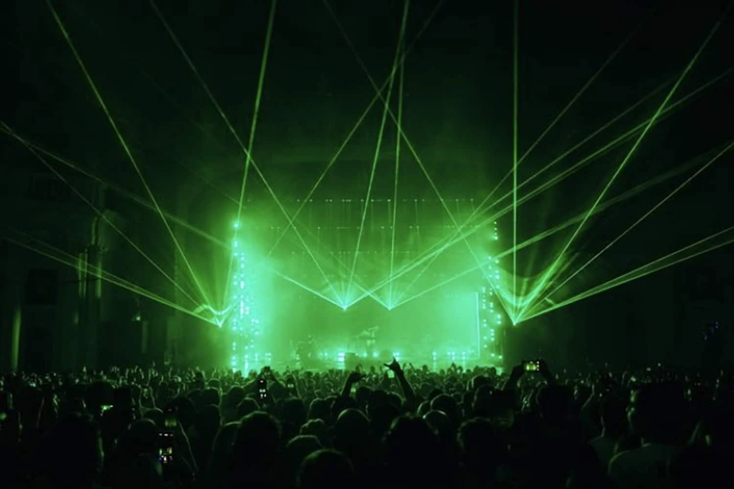 A large crowd at a concert or music event with green laser lights illuminating the stage and audience.
