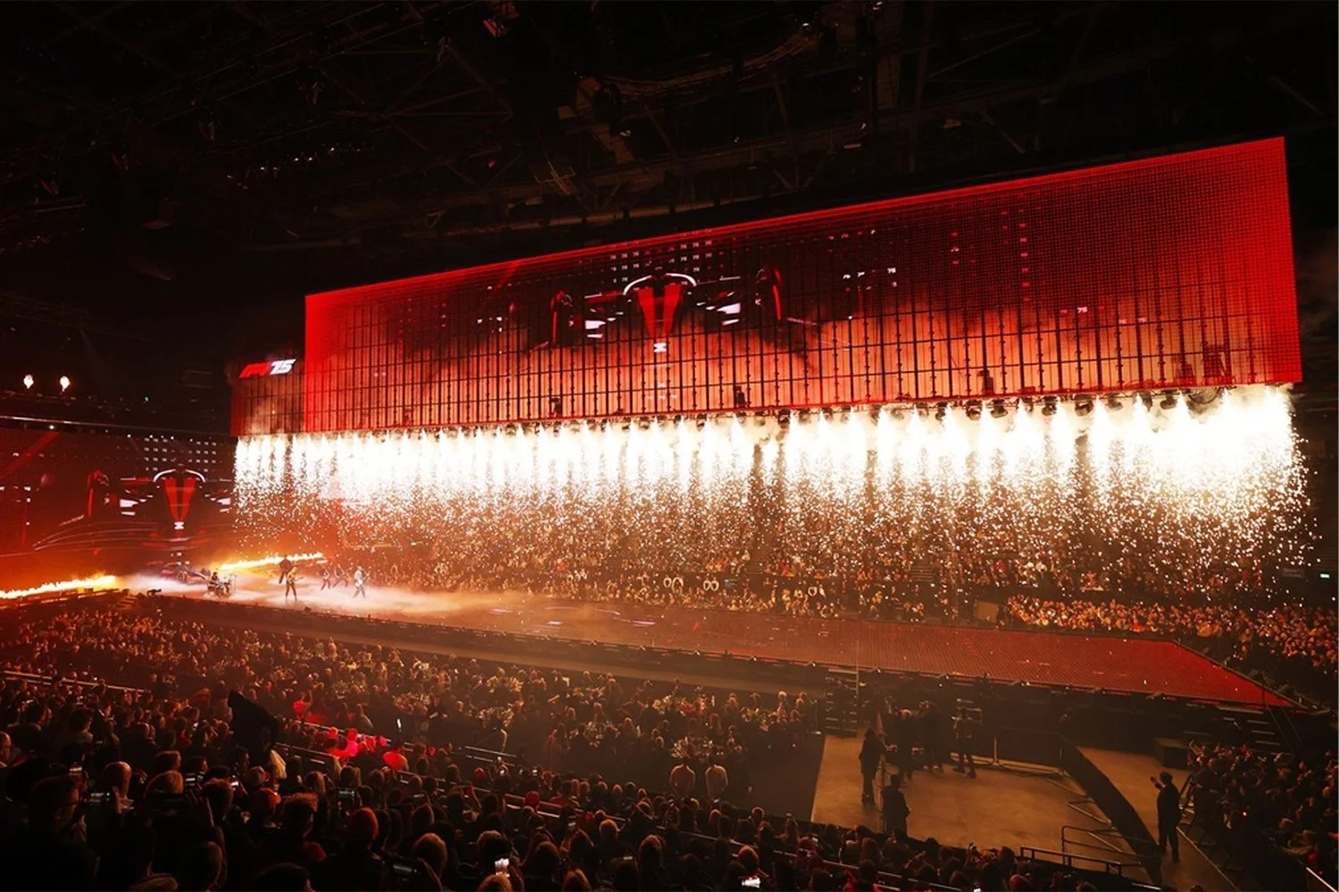A large indoor concert or event with a crowd in front of a stage. The stage features a red LED screen display and pyrotechnics shooting upward. The scene is vibrant and energetic with a spectacle of lights and smoke effects.