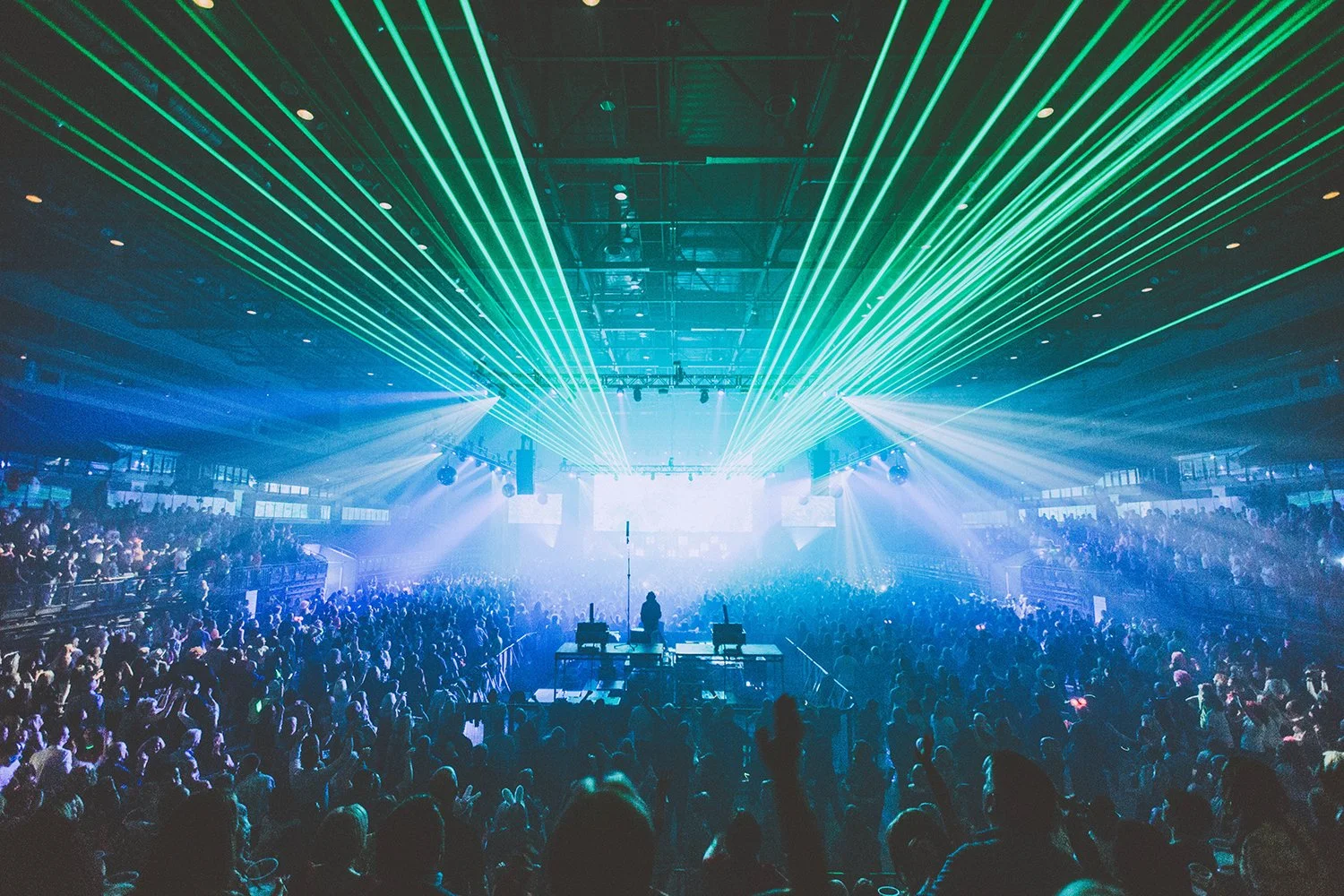 A large indoor concert or music festival with a crowd of people, colorful laser lights, and a stage at the front with performers.