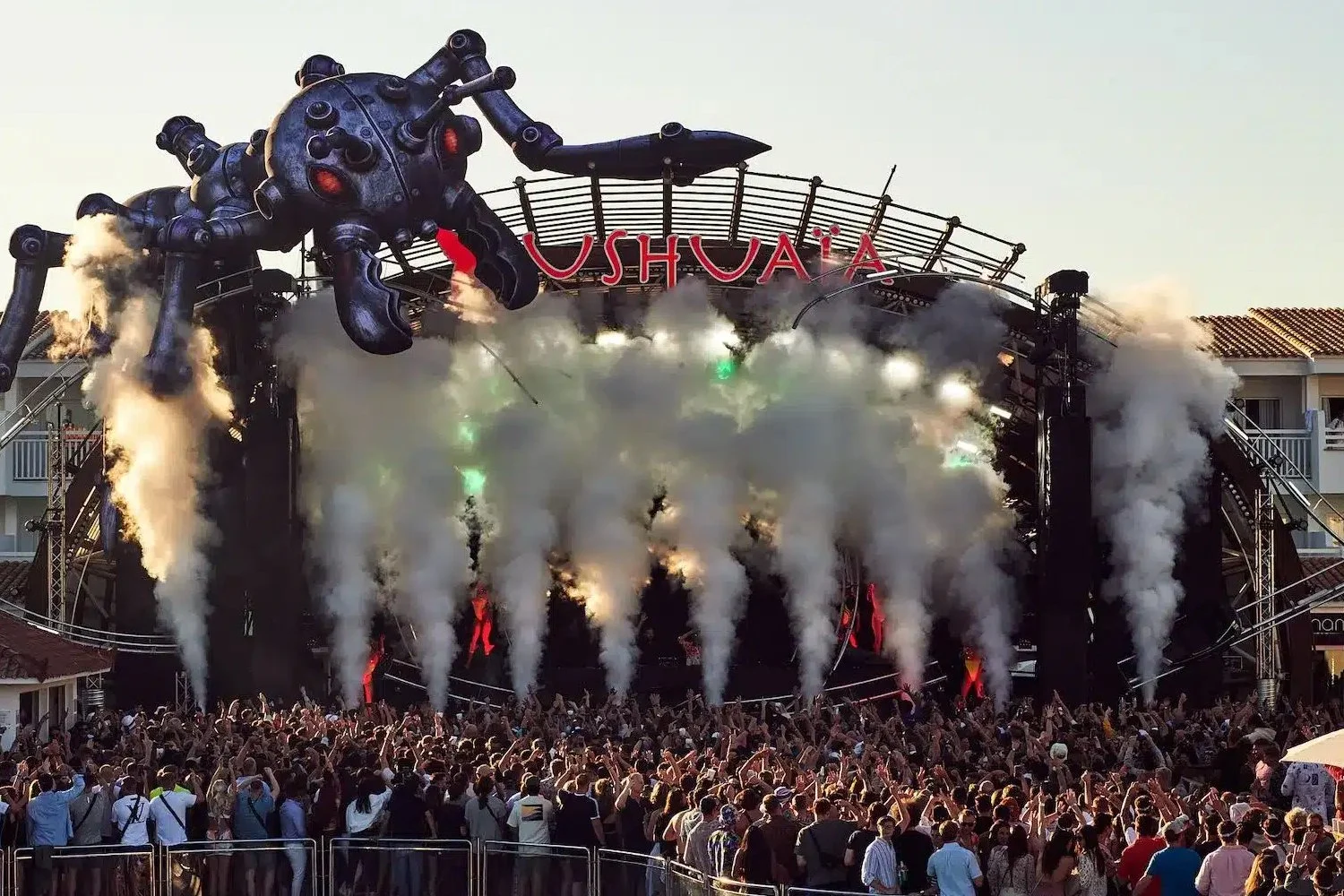 Large outdoor concert stage with smoke and fog effects, a giant spider-like sculpture above, and a crowd of people dancing and enjoying the show at sunset.