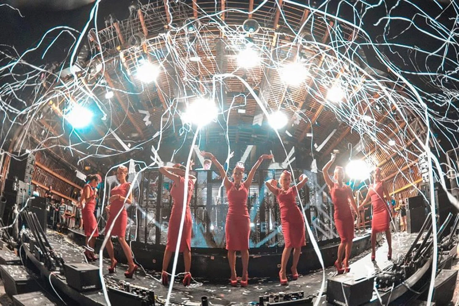 A group of women in red dresses and high heels dancing on a stage with bright lights, confetti, and streamers, in a lively party or club setting.
