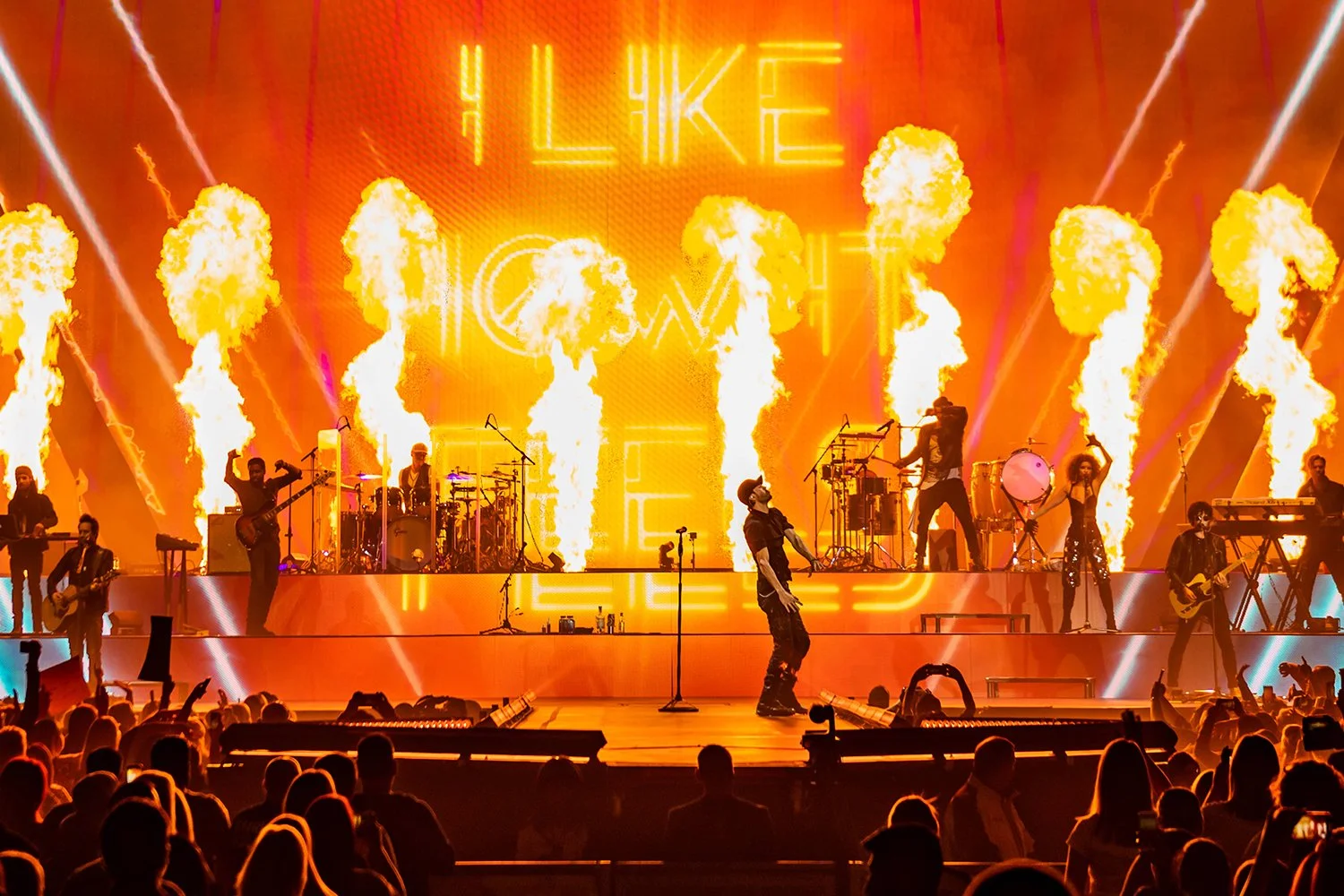 A live concert scene with a band performing on stage surrounded by flames and laser lights in an indoor arena