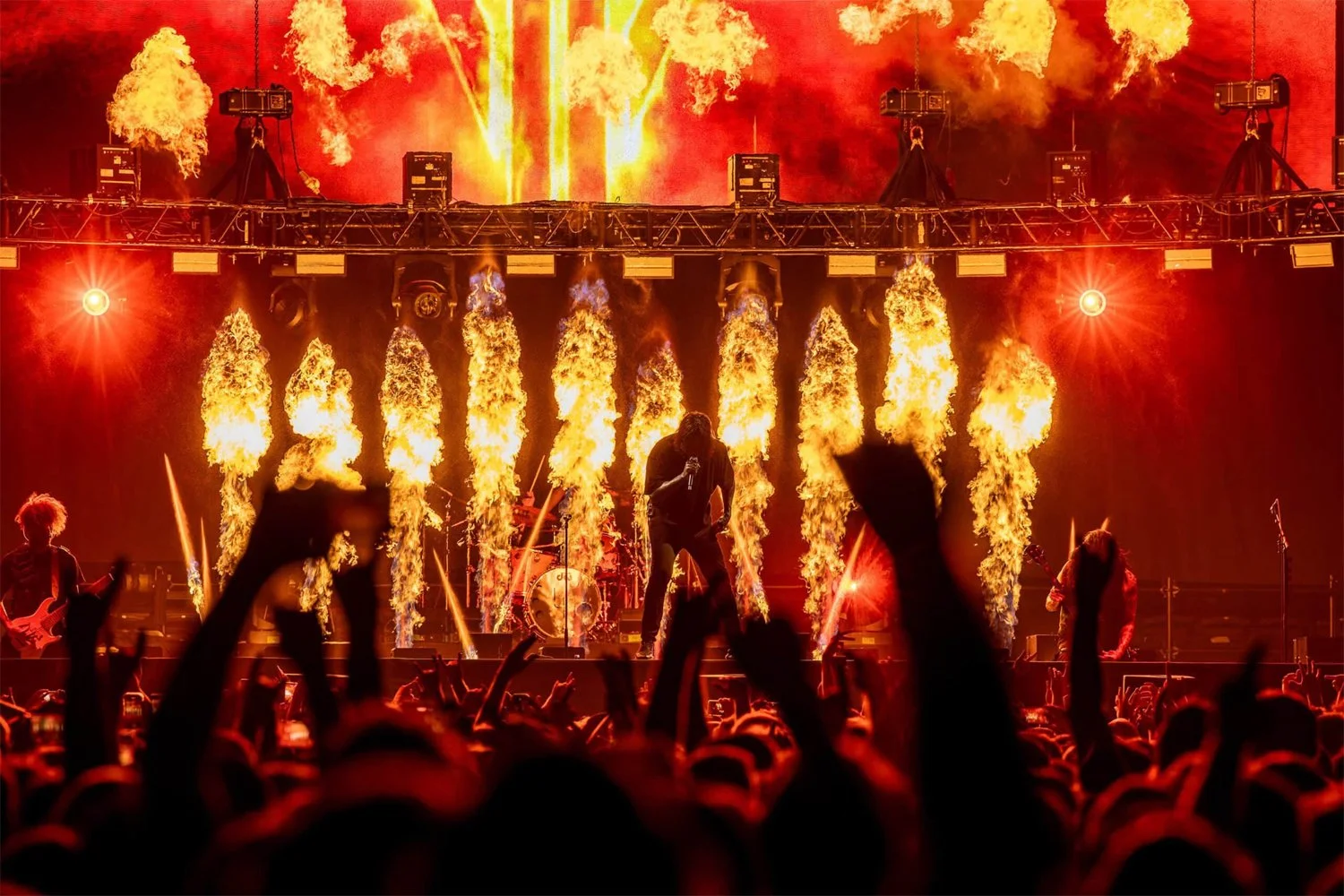 A concert stage with flames and fire effects, with a band performing amidst pyrotechnics and an audience with raised hands.