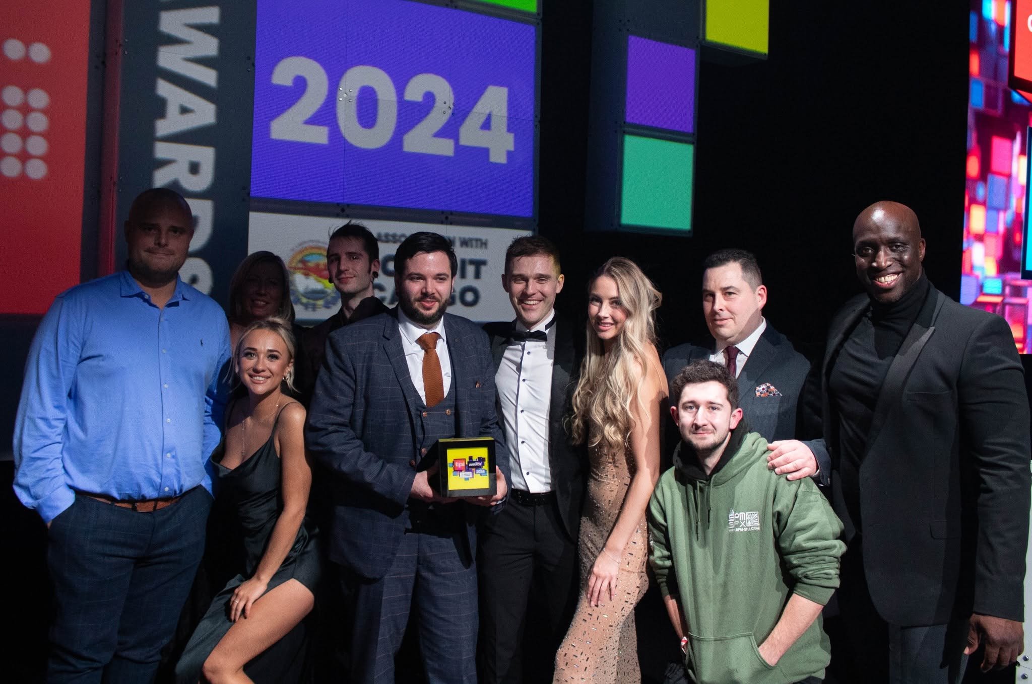 A group of people dressed in formal and casual attire posing together on a stage or at an event, with a large screen in the background displaying the year '2024' and other graphics.