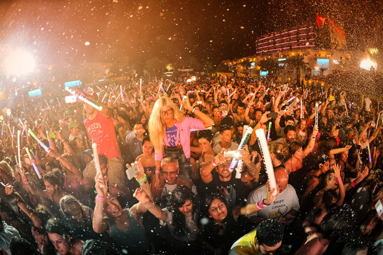 Crowd at a lively concert or festival at night, with people holding glow sticks and taking photos, colorful lights, and water spray in the air.