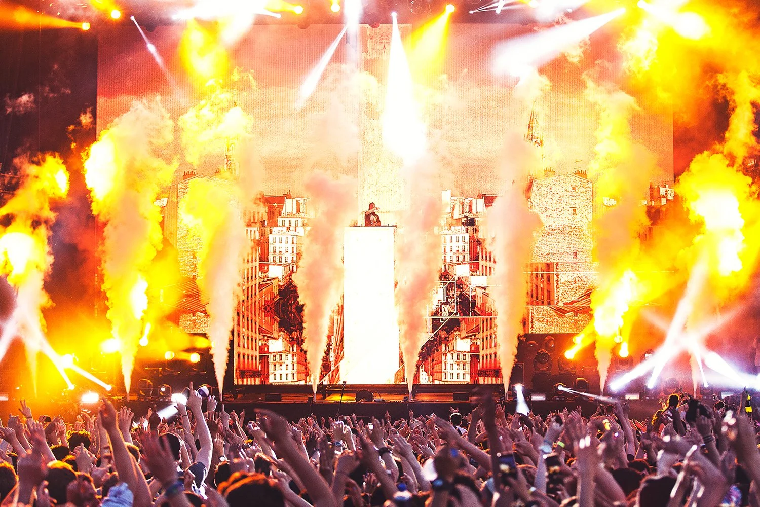 Crowd at a concert with DJ on stage, surrounded by bright yellow and orange stage lights and smoke effects, with a cityscape backdrop.