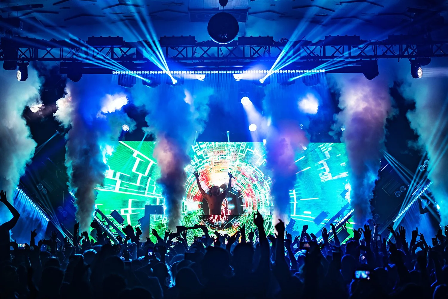 A lively concert scene with a DJ or performer on stage, illuminated by vibrant blue and green lights and digital screens, with a crowd dancing and raising their hands.