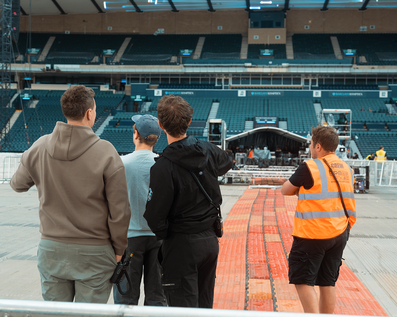 Four crew members standing on a concert stage in an empty stadium, looking towards the stage setup with equipment and scaffolding.
