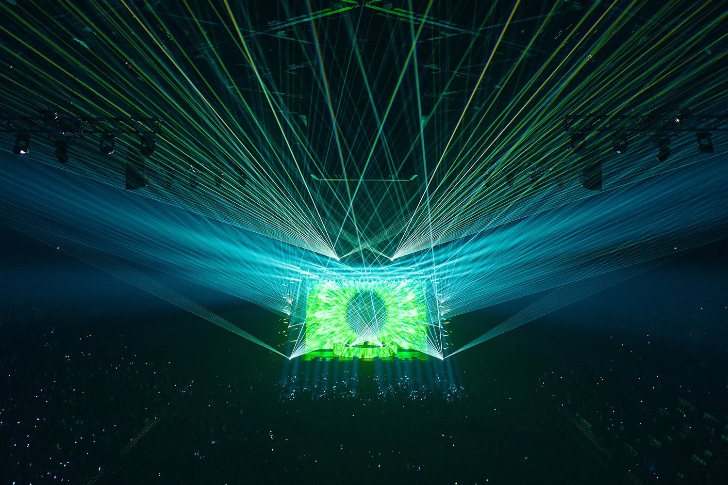 A concert stage with a DJ, surrounded by green and white laser lights projecting in various directions, creating an energetic atmosphere.