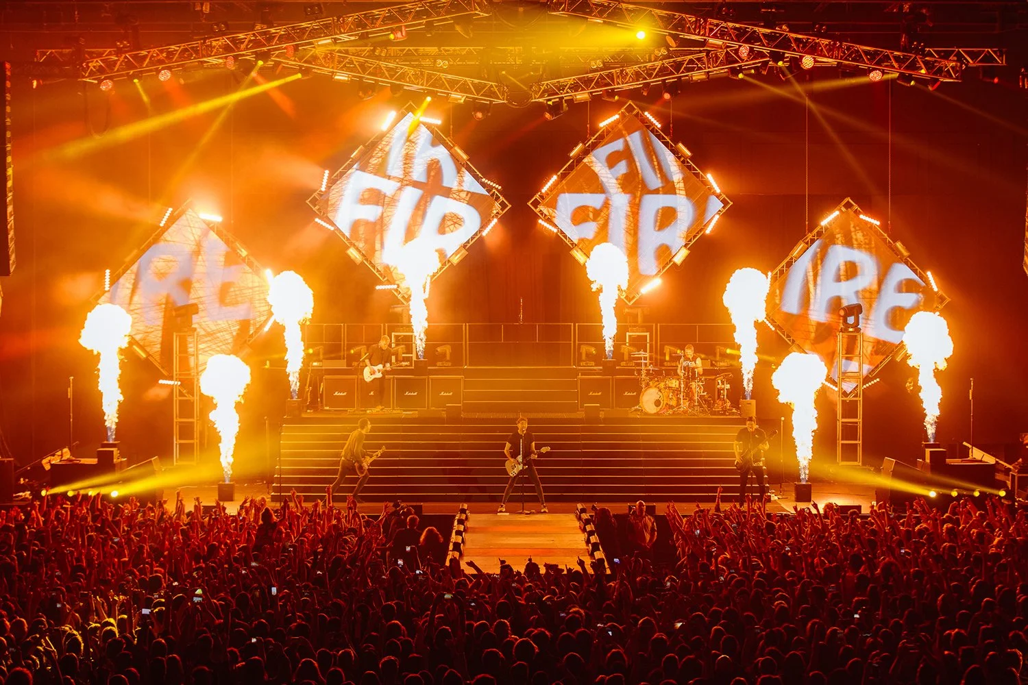 A live concert with a band performing on stage, illuminated by bright yellow and orange lights, with large screens displaying the word "FUEL" in the background, fireworks, and an enthusiastic crowd in the foreground.