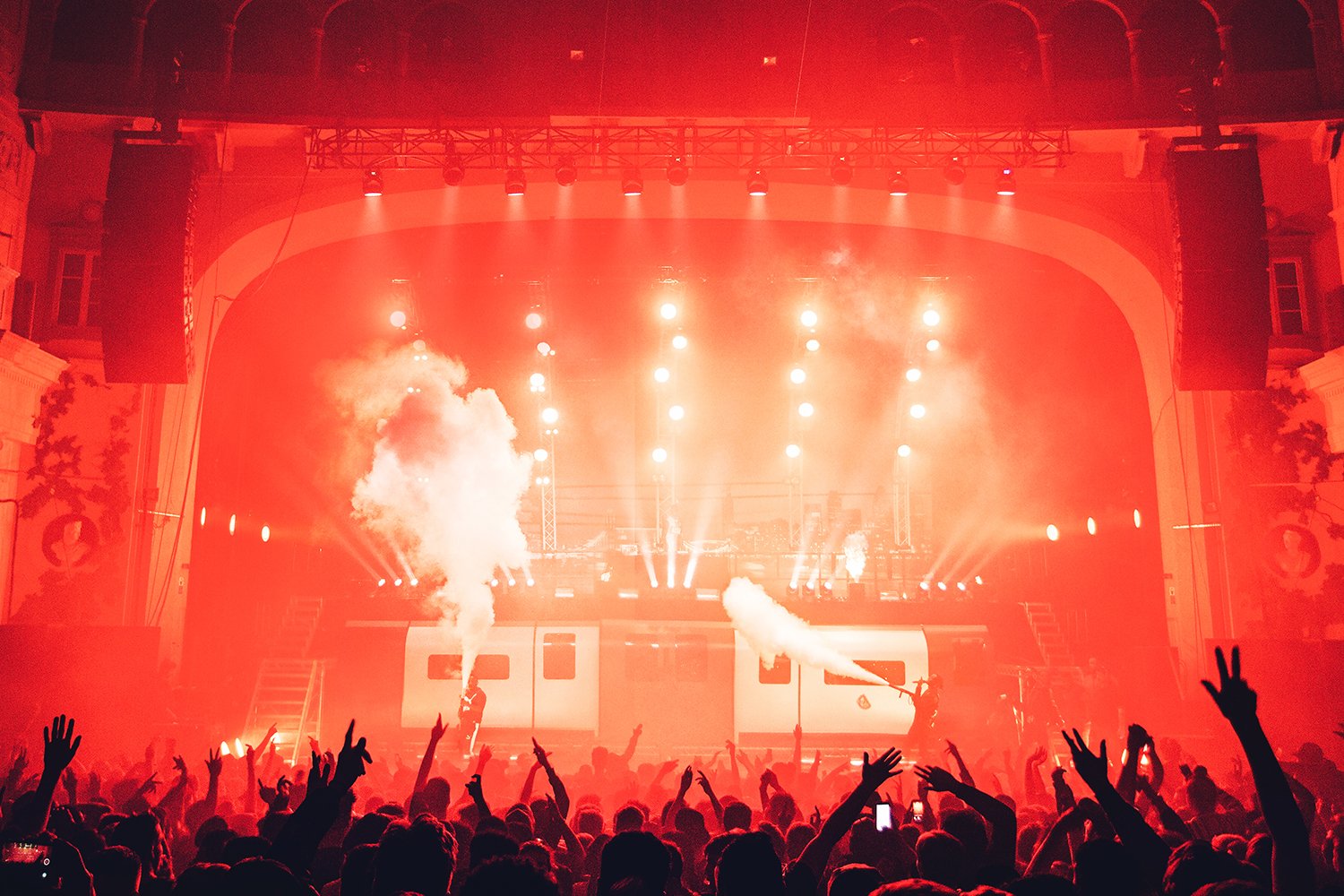 Concert stage with bright lights and smoke effects, crowd with hands raised, indoors with ornate architecture.